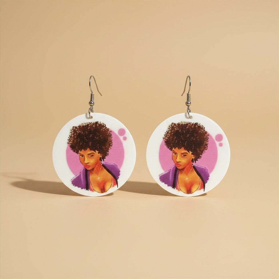 Close-up of round white disc earrings featuring a printed illustration of a woman with an afro in a purple top against a pink background, on a beige textured backdrop.