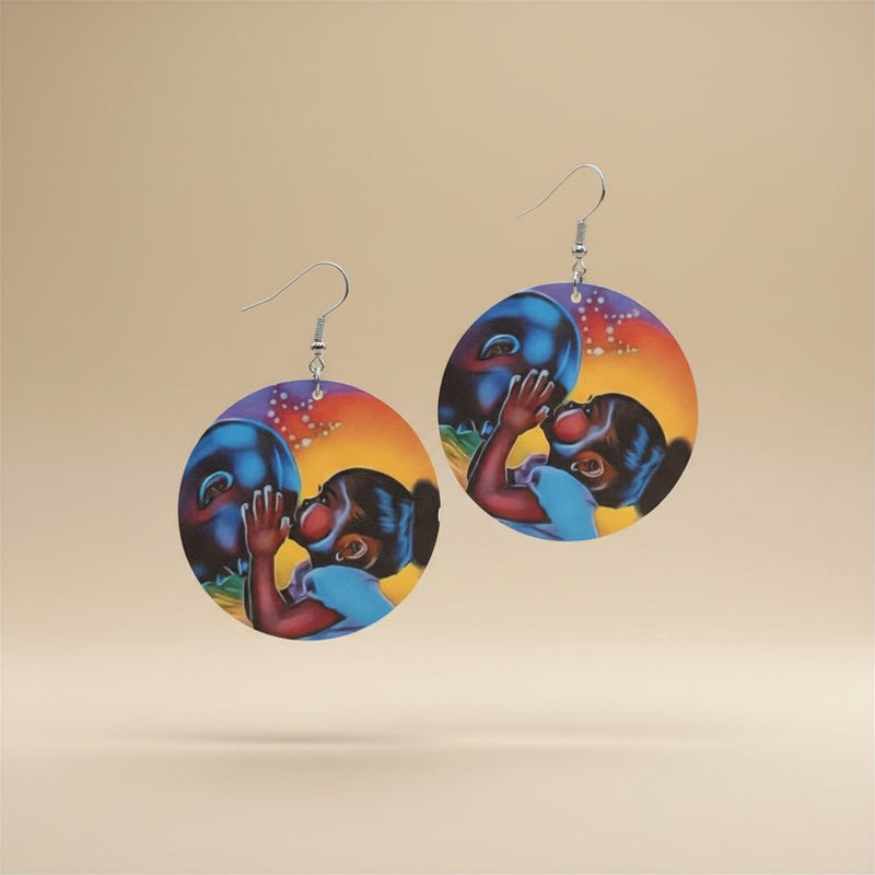 Flat lay of the Mother and Child African Art Earrings — round dangle earrings printed with a colourful figure illustration in blue, orange, red and purple tones, on a plain beige background.