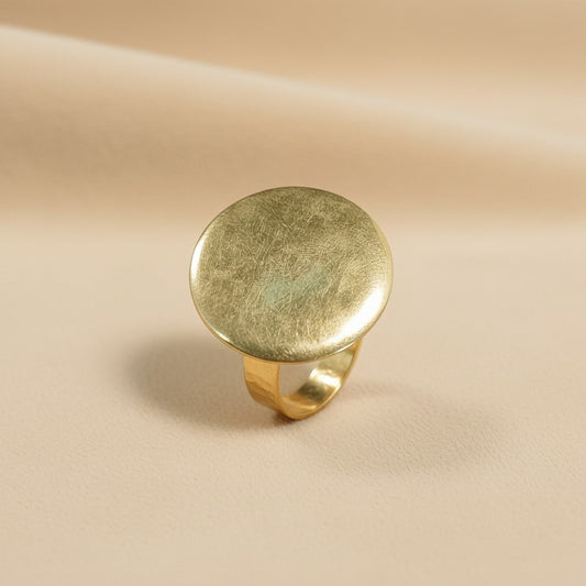Tinkers brushed gold-tone medallion ring with a large circular face on a soft beige fabric background.