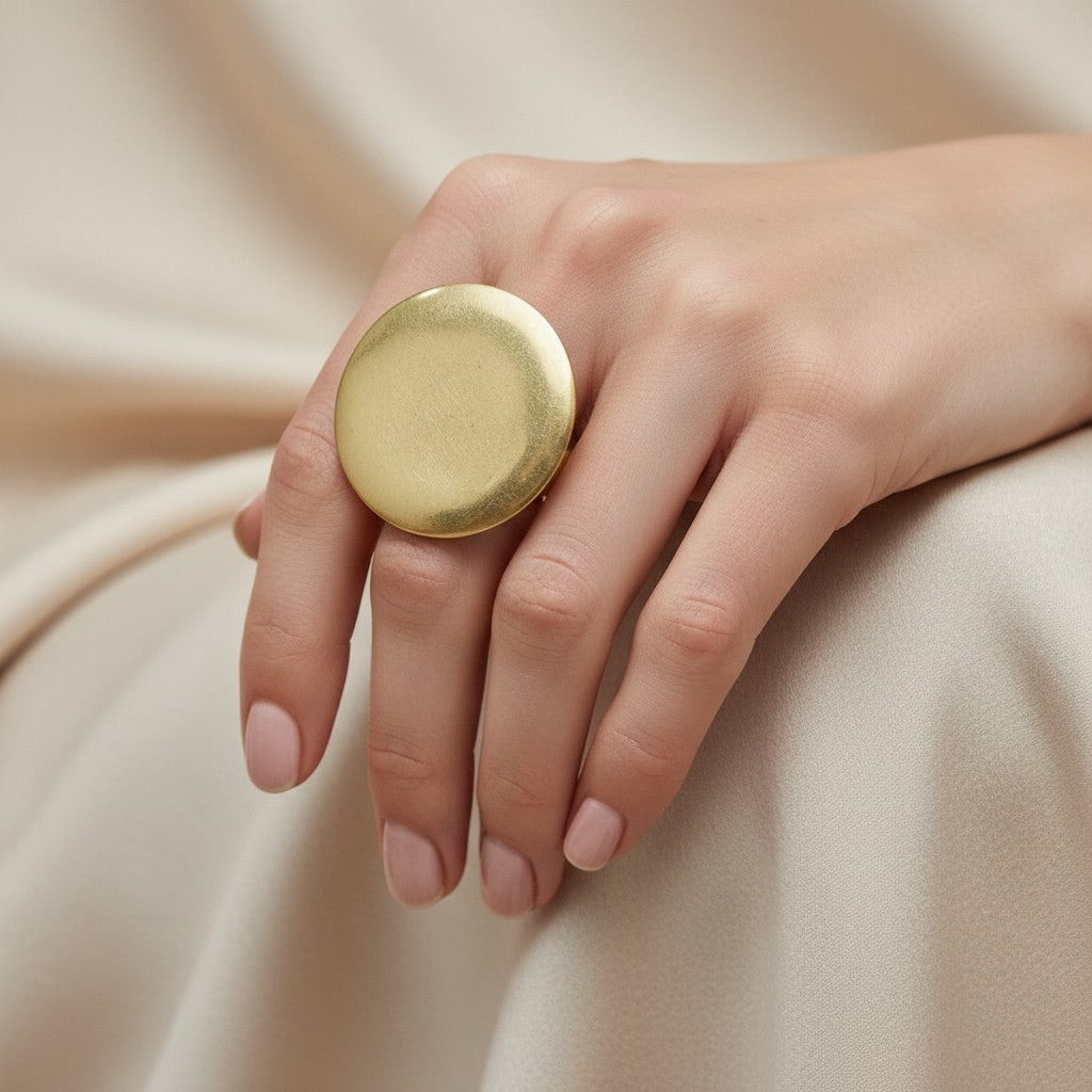 Tinkers gold-tone brushed metal medallion ring with a large circular face worn on a hand against beige fabric.