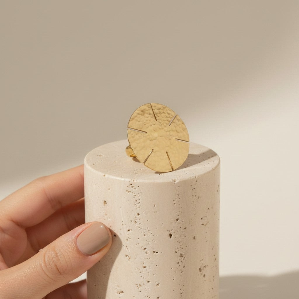 Tinkers hammered brass disc statement ring with sun-ray cuts on a textured surface, displayed on a stone pedestal.
