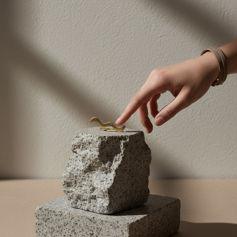 Tinkers handmade raw brass wrap ring with a wavy S-curve design resting on a stone pedestal with a hand reaching out.